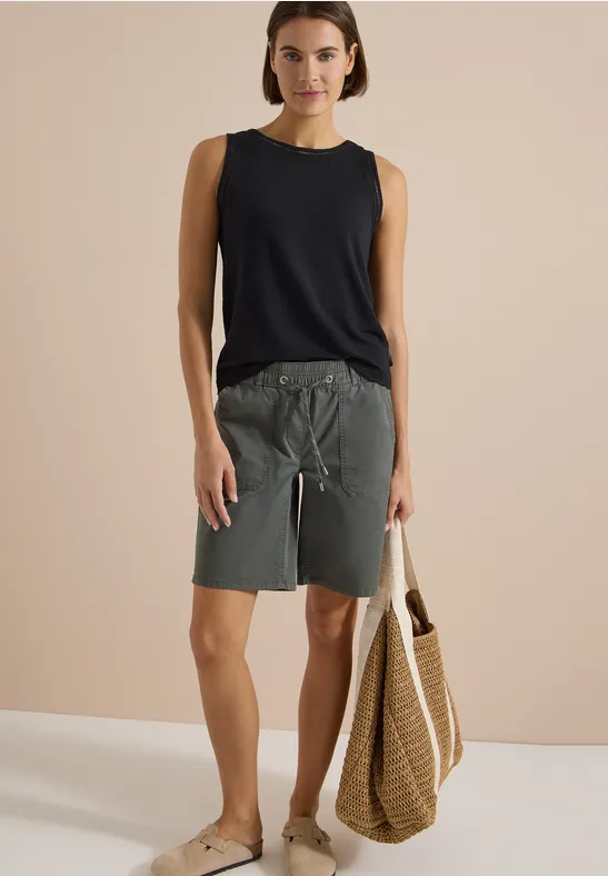 Frau in ärmellosem schwarzen Top, grauen Shorts und Sandalen, trägt eine geflochtene Strandtasche.