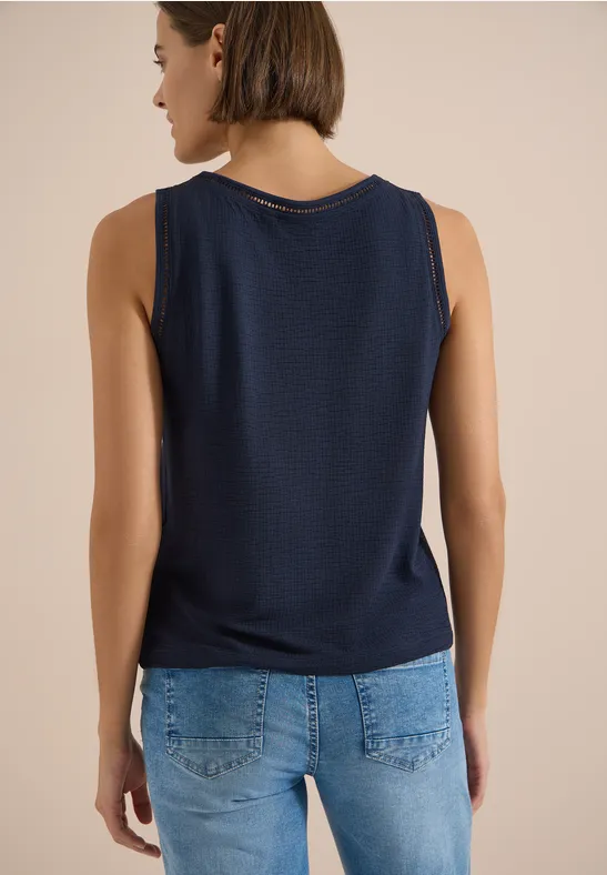 Frau von hinten in ärmellosem dunkelblauem Top und blauen Jeans vor neutralem Hintergrund.