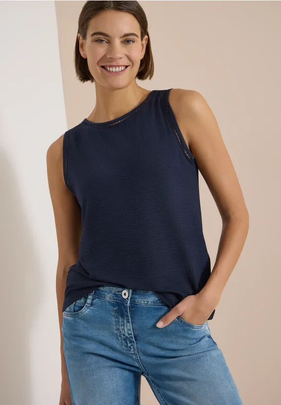 Frau in ärmellosem dunkelblauem Top und blauen Jeans, Hand in Tasche, vor beigem Hintergrund.
