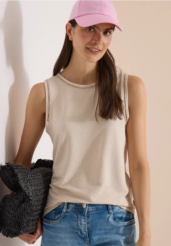 Vrouw in beige tanktop, blauwe jeans en roze pet, met gevlochten tas voor een lichte achtergrond.