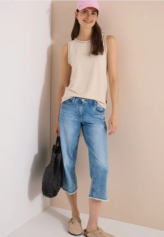 Vrouw in beige mouwloos topje, lichtblauwe capri-jeans en sandalen, met een zwarte tas voor een muur.