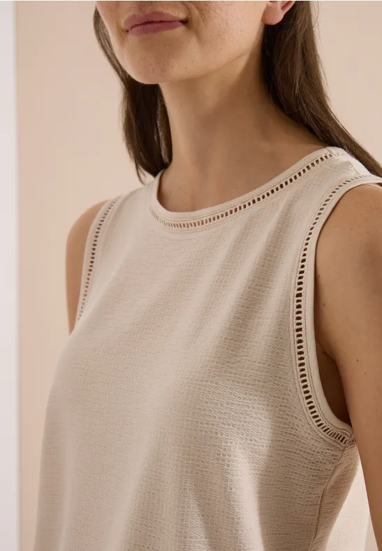 Close-up van een vrouw in een mouwloze beige top met ajourwerk langs de rand.
