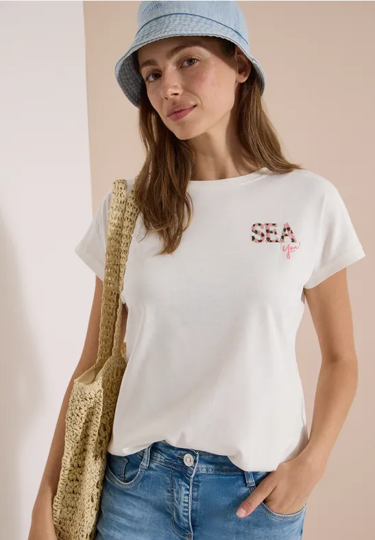 Frau trägt weißen SEA-T-Shirt, Jeans, Strohhut und gehäkelte Umhängetasche, steht mit Hand in der Hosentasche.