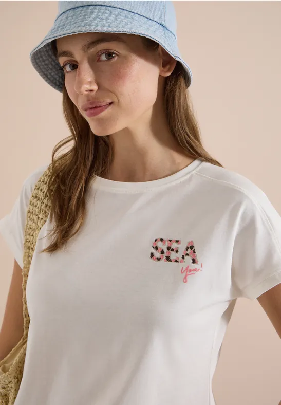 Frau mit hellblauem Fischerhut, weißem T-Shirt mit "SEA you"-Aufdruck und Strohtasche.