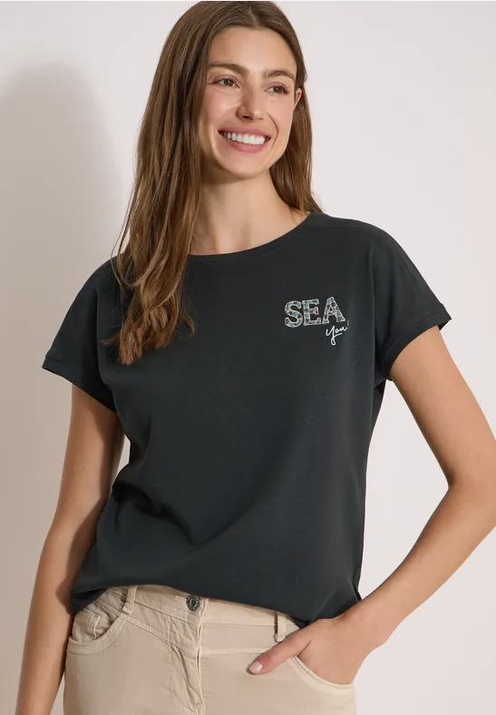 Frau in schwarzem 'SEA'-T-Shirt und beigen Hosen, lässig stehend mit einer Hand in der Tasche.