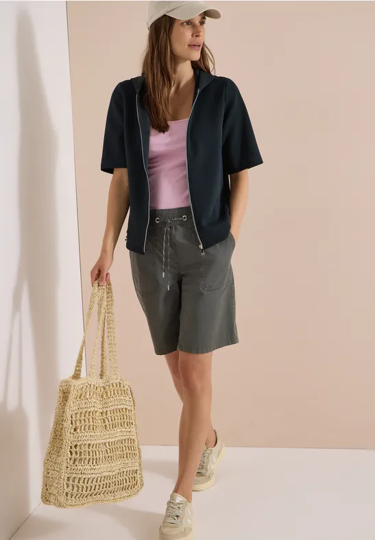 Frau trägt graue Bermudashorts, helles Top, dunkelblaues Hemd, Mütze, Strohtasche und Sneaker.