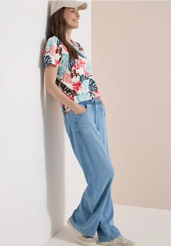 Eine Person lehnt entspannt an einer Wand, trägt ein buntes Blumen-T-Shirt, weite Jeans und weiße Sneaker.