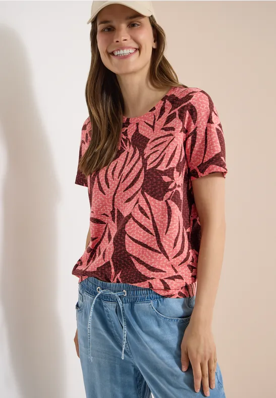 Frau im kurzärmligen, korallrosa Shirt mit tropischen Blattmotiven und hellblauen Jeans-Shorts.