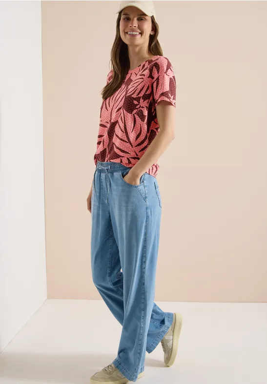 Frau mit pinkem Blattmuster-T-Shirt und lockeren Jeans vor neutralem Hintergrund.