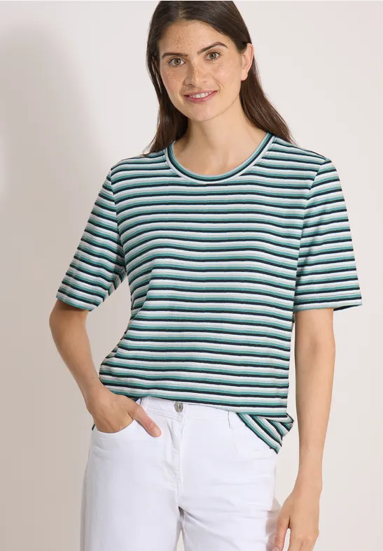 Eine Frau trägt ein kurzärmeliges gestreiftes T-Shirt in Blau, Grün und Weiß und weiße Jeans.