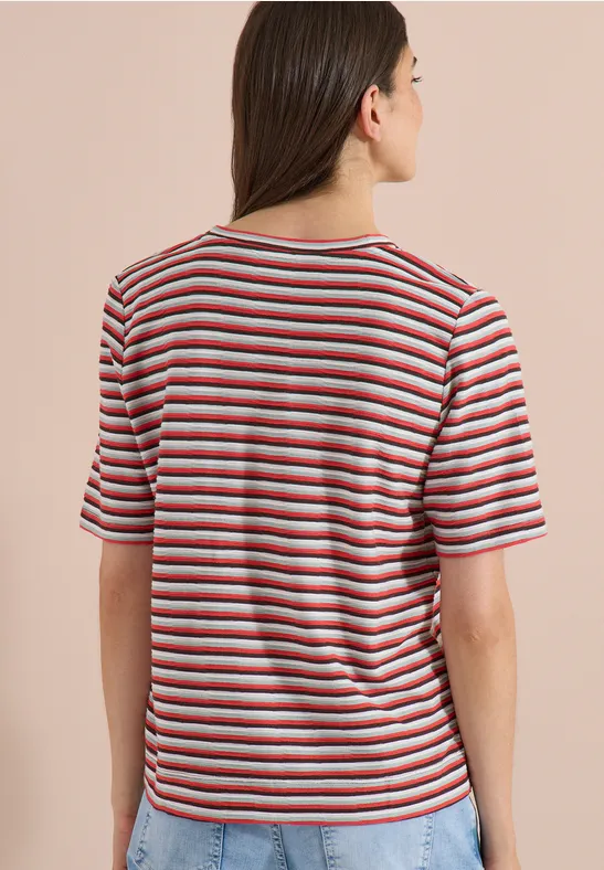 Frau von hinten, trägt gestreiftes T-Shirt mit roten, weißen und blauen Streifen vor beigem Hintergrund.