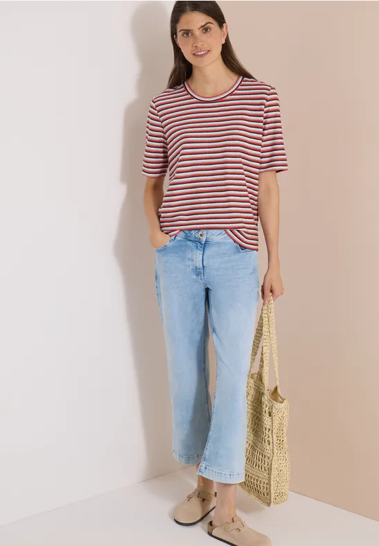 Frau trägt gestreiftes T-Shirt, helle Jeans und hält eine geflochtene Strohtasche, steht vor heller Wand.