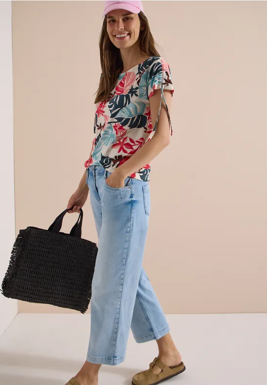 Femme en top à fleurs, jean large clair, sandales et sac panier noir sur fond beige.