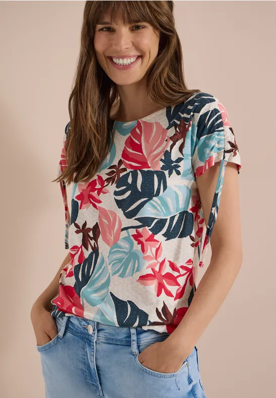 Femme en jeans et t-shirt clair orné de feuilles tropicales colorées, mains dans les poches.