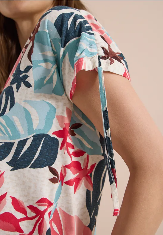 Femme porte un top blanc à manches courtes orné de feuilles turquoise-noir et de fleurs rouges, avec un lien sur la manche.