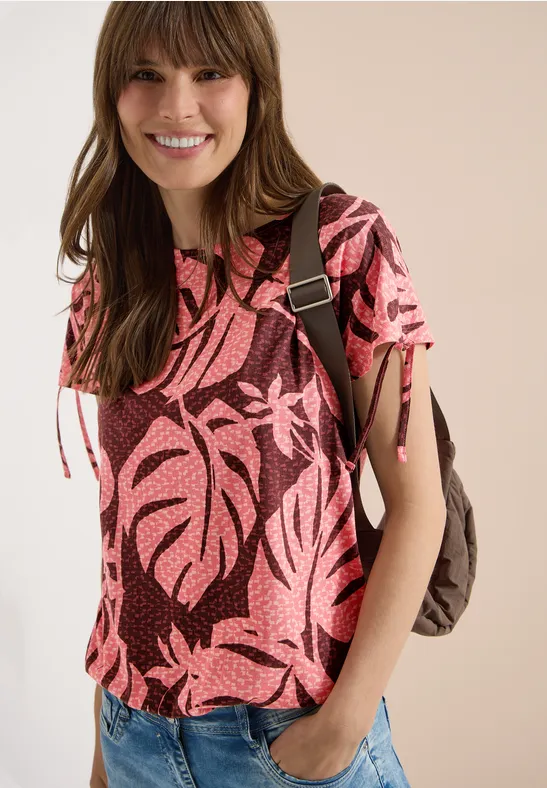 Frau mit rosa Blattmuster-Shirt, Jeans und Rucksack vor hellem Hintergrund.