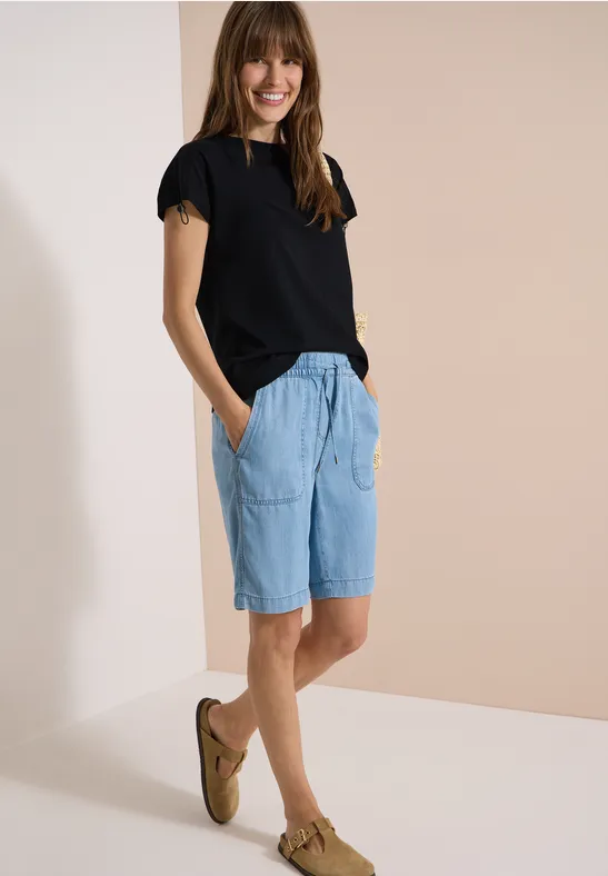 Frau in schwarzem T-Shirt, hellblauen Jeans-Shorts und braunen Slippern vor schlichtem, hellem Hintergrund.