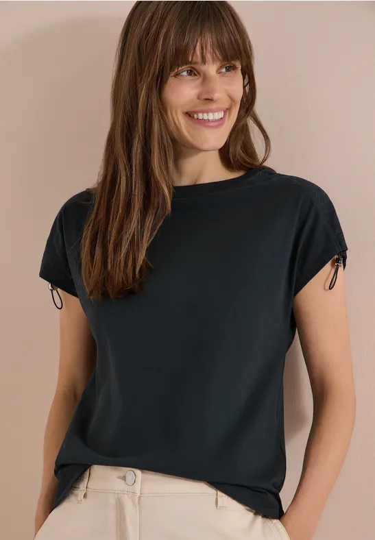 Eine Frau trägt ein schwarzes T-Shirt und eine beigefarbene Hose vor neutralem Hintergrund.