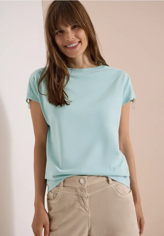 Frau trägt hellblaues T-Shirt und beige Hose vor hellem Hintergrund.