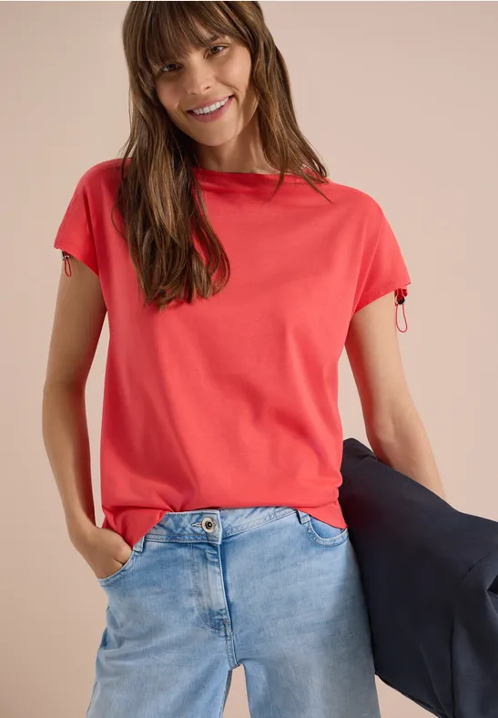 Vrouw draagt rood T-shirt, blauwe spijkerbroek en houdt een zwarte jas vast.
