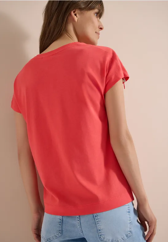 Vrouw van achteren in een koraalkleurig t-shirt en lichtblauwe spijkerbroek voor een neutrale achtergrond.