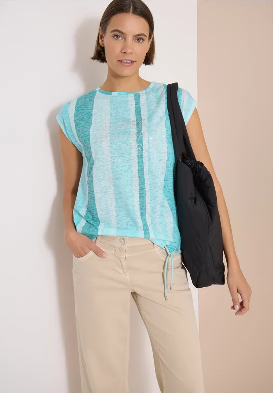 Femme en haut rayé turquoise et blanc, pantalon beige et sac noir, pose décontractée.