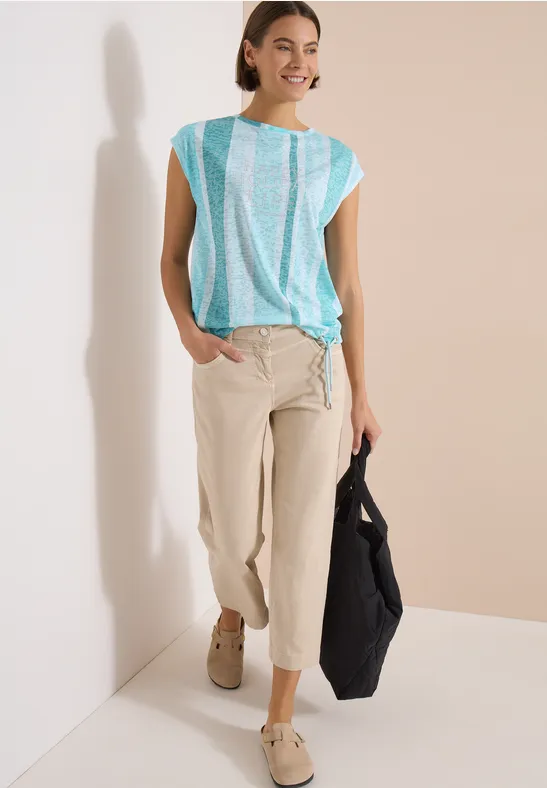 Personne en blouse turquoise à rayures blanches et pantalon beige, tenant un sac noir.