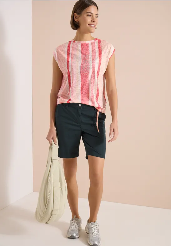 Femme en top rayé rose et blanc, bermudas noirs, baskets grises et sac vert clair.