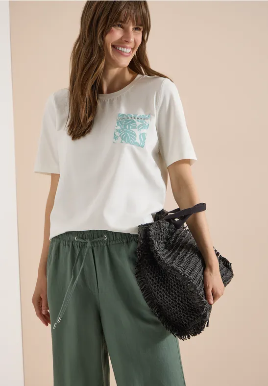 Frau trägt weißes T-Shirt mit türkisem Brusttaschen-Print, weite grüne Hose und großen schwarzen Netzbeutel.