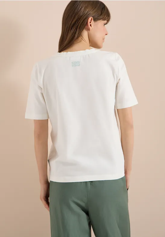 Frau von hinten, trägt weißes T-Shirt und grüne Hose vor beigem Hintergrund.