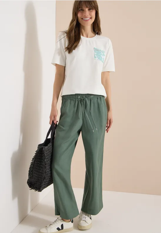 Frau trägt weißes T-Shirt mit gemusterter Tasche, weite grüne Hose, weiße Sneaker und schwarzen Netzbeutel.
