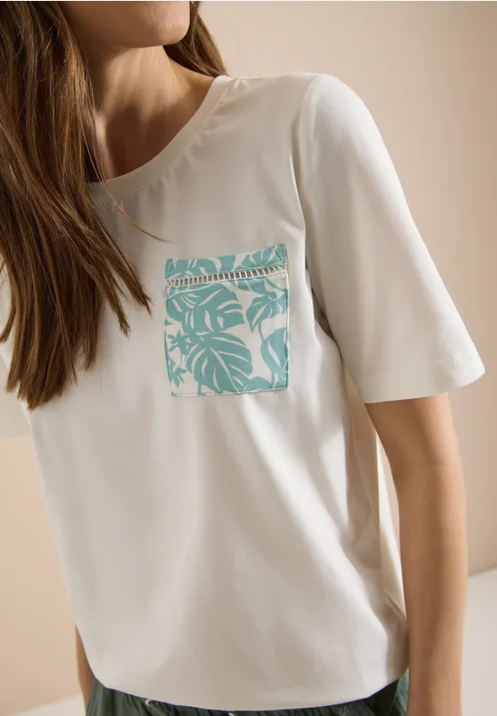 Weißes T-Shirt mit türkisem tropischem Blattmuster auf der Brusttasche, getragen von einer Frau.