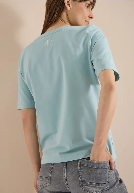 Person von hinten mit hellblauem T-Shirt und grauen Jeans vor neutralem Hintergrund.