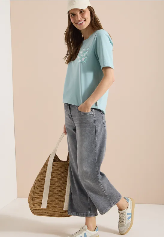 Frau in lockerem pastellblauen T-Shirt, weiten Jeans-Culottes, Strohtasche und Sneakern.