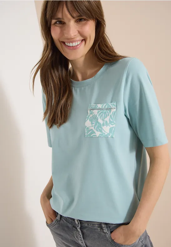 Eine junge Frau trägt ein hellblaues T-Shirt mit kurzem Arm und gemustertem Brusttaschen-Print.