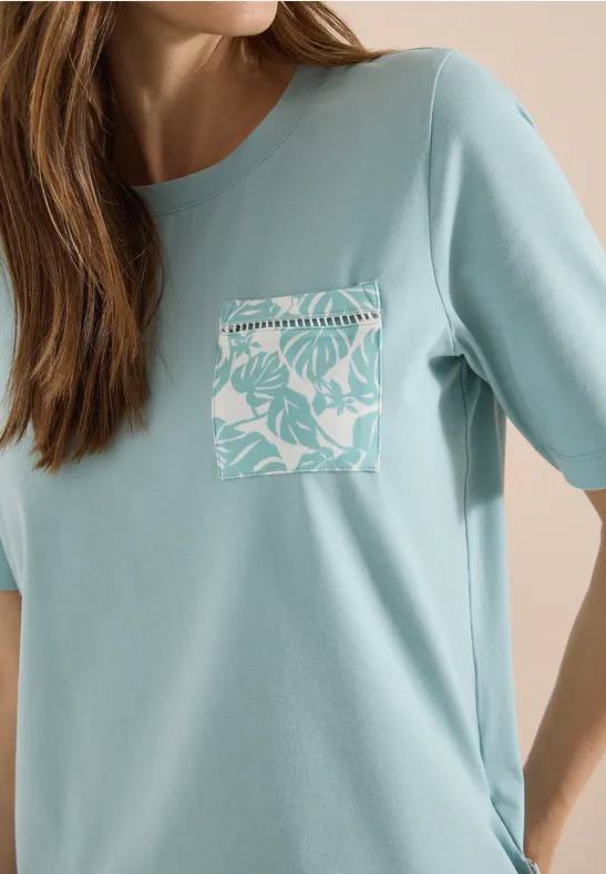 Frau trägt hellblaues T-Shirt mit aufgesetzter Brusttasche in Weiß mit tropischem Blattmuster.