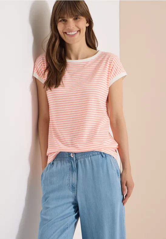 Frau im gestreiften T-Shirt und leichter Jeanshose lehnt an hellem Hintergrund.