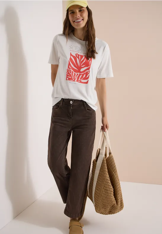 Frau in weißem T-Shirt mit rotem Print, braunen Jeans und großer Strohtasche vor hellem Hintergrund.