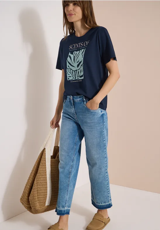 Frau trägt dunkelblaues T-Shirt mit Aufdruck, lockere Jeans und eine große Basttasche.