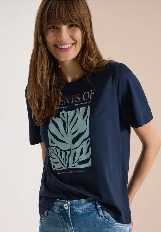 Frau in dunkelblauem T-Shirt mit grünem Blattmuster und blauen Jeans.