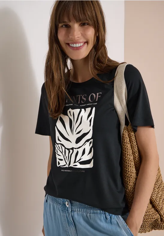 Frau vor neutralem Hintergrund, trägt schwarzes T-Shirt mit weißem Zebramuster, blaue Jeans, Strohtasche.