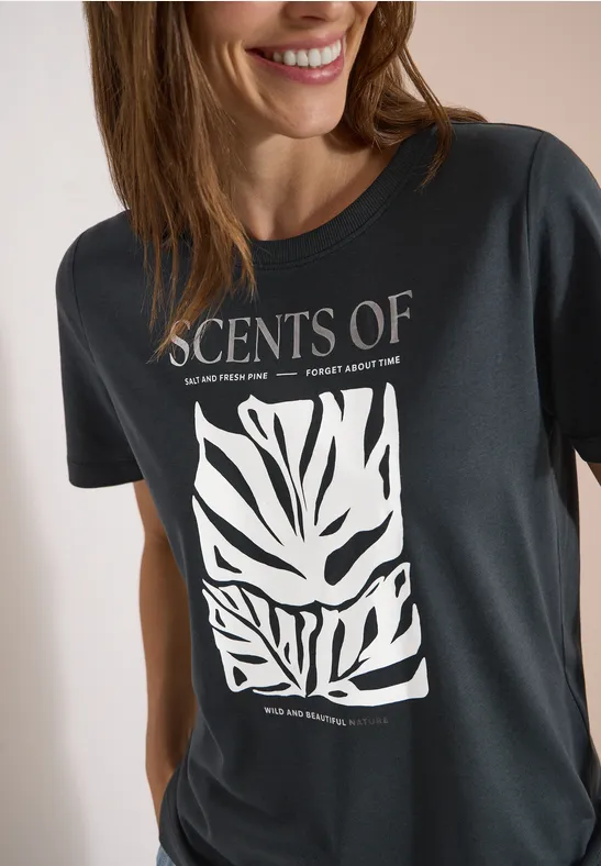 Frau in dunklem T-Shirt mit Aufdruck 'SCENTS OF' und weißem, abstraktem Blattmuster.
