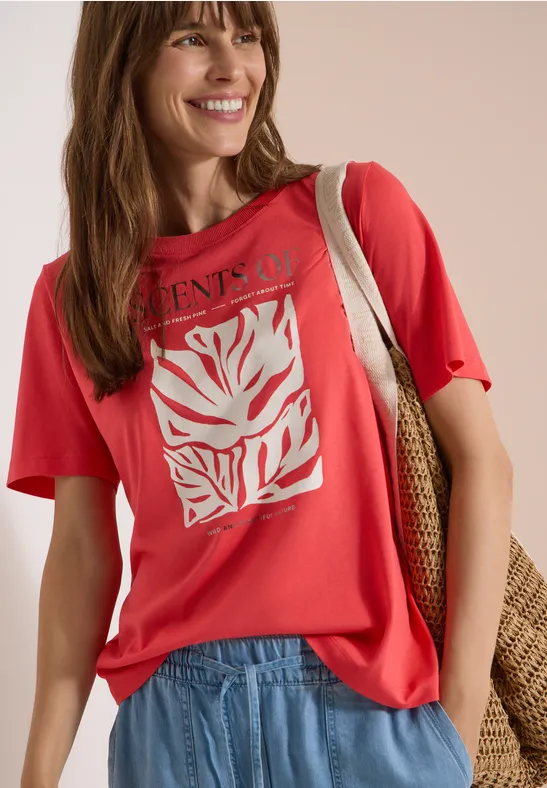 Frau trägt rotes T-Shirt mit weißem Blatt-Print, hellblaue Jeans und Basttasche über der Schulter.