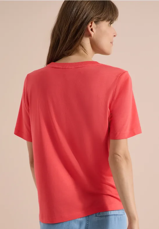 Frau steht mit dem Rücken zur Kamera, trägt rotes T-Shirt und Jeans vor hellem Hintergrund.
