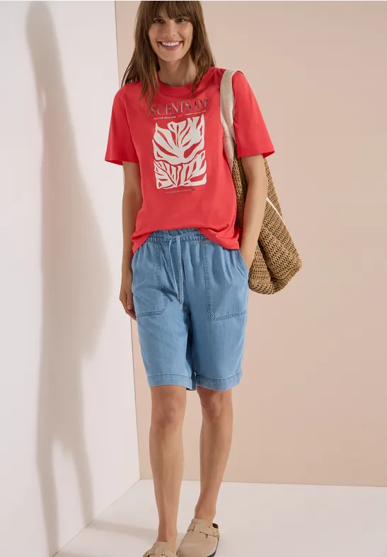 Eine Frau trägt rotes T-Shirt mit weißem Motiv, lockere hellblaue Shorts und eine Korbtasche.