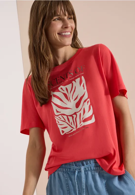 Frau mit rotem T-Shirt mit weißem grafischen Motiv und blauer Jeans, stehend vor neutralem Hintergrund.