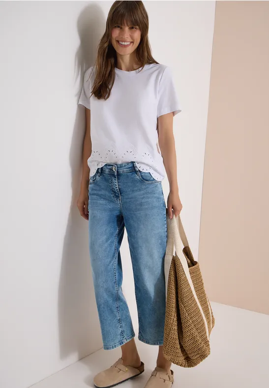 Frau in weißem T-Shirt mit Spitzensaum, weitem Jeans, Slip-on-Schuhen und Strohtasche vor hellem Hintergrund.