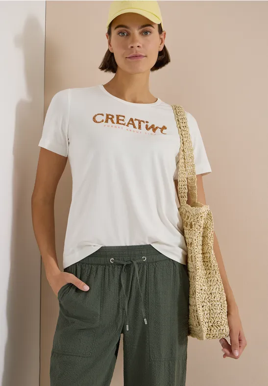 Person trägt weißes T-Shirt mit 'CREATIVE'-Aufdruck, grüne Hose und gehäkelte Tasche.