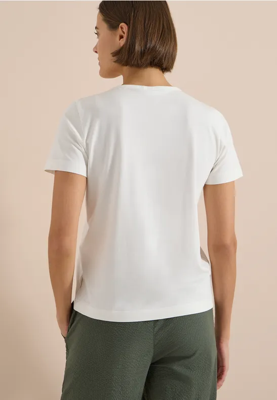 Rückansicht einer Person in weißem T-Shirt und grüner Hose vor beigem Hintergrund.