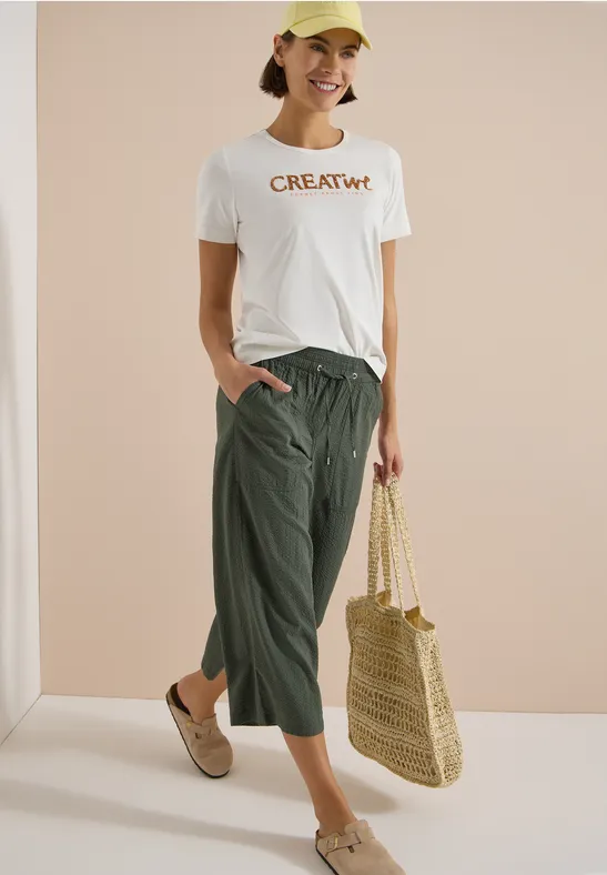 Frau trägt weißes T-Shirt mit 'CREATIVE'-Aufdruck, grüne Culotte, Strohtasche und beigen Mules vor neutralem Hintergrund.